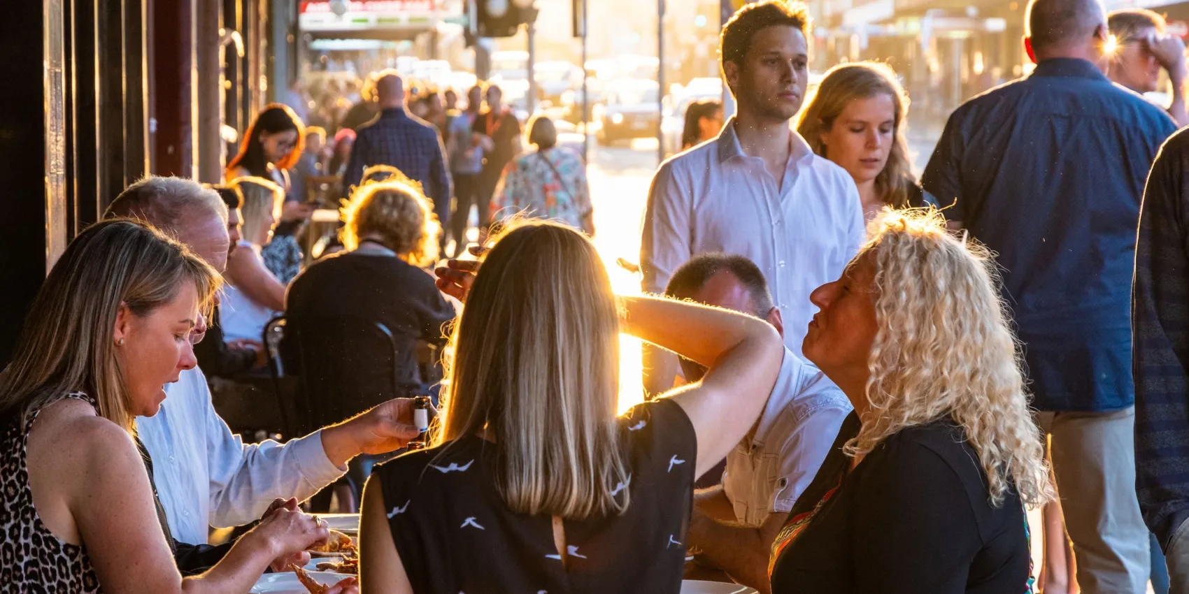 Busy outdoor dining area at sunset, with people eating and walking along a crowded city street in warm golden light.