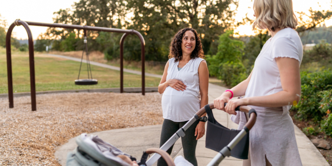 An eight‑months‑pregnant woman chats with a friend pushing a baby stroller.