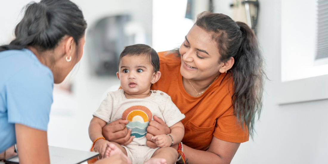 Caregiver holding an infant while speaking with a healthcare worker during a child health and immunisation appointment.